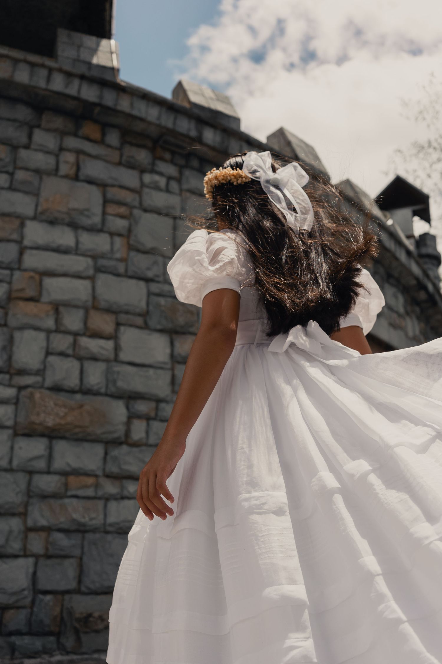 A flowing white christening dress billows in the wind against a stone wall backdrop on a cloudy day.