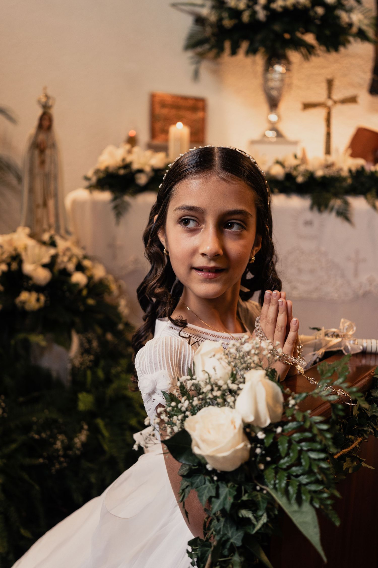 A young flower girl in a white dress poses with roses and greenery in a romantic indoor setting.