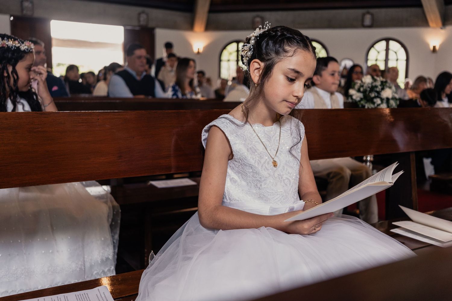 A person in a white dress reads a document while seated in a wooden pew at a formal event.