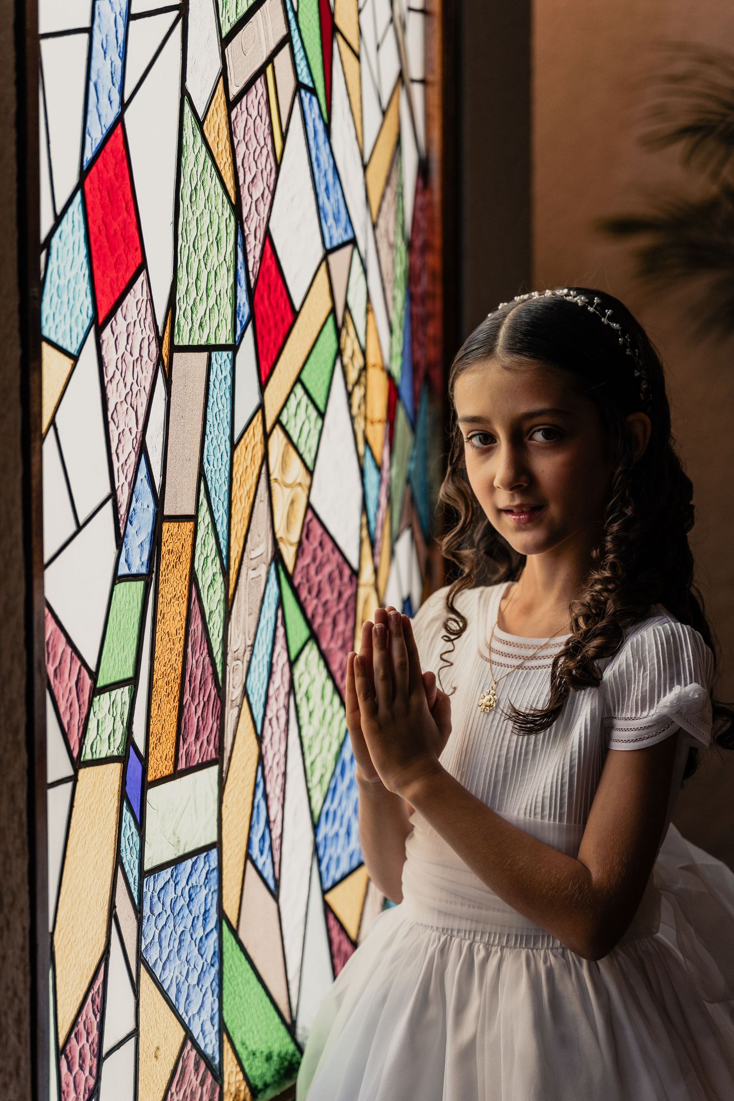 Young girl in white dress poses by colorful stained glass windows in artistic lighting.