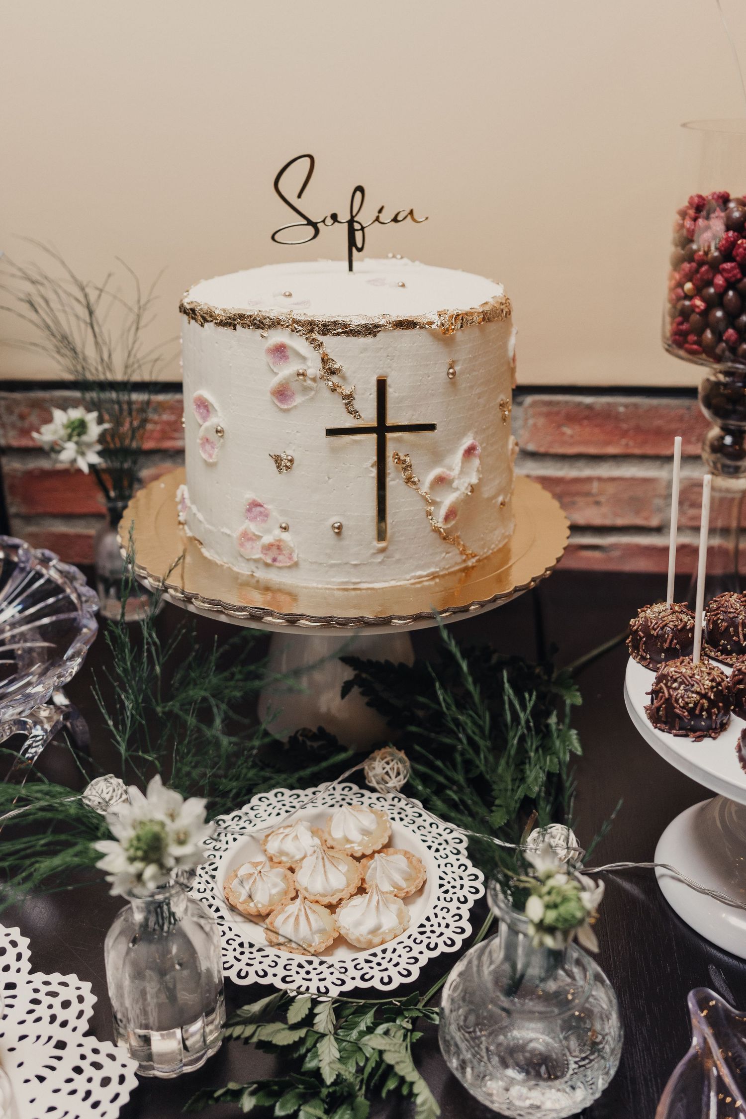 Elegant dessert table with white cake, macarons and decorative doilies on rustic display with greenery accents.