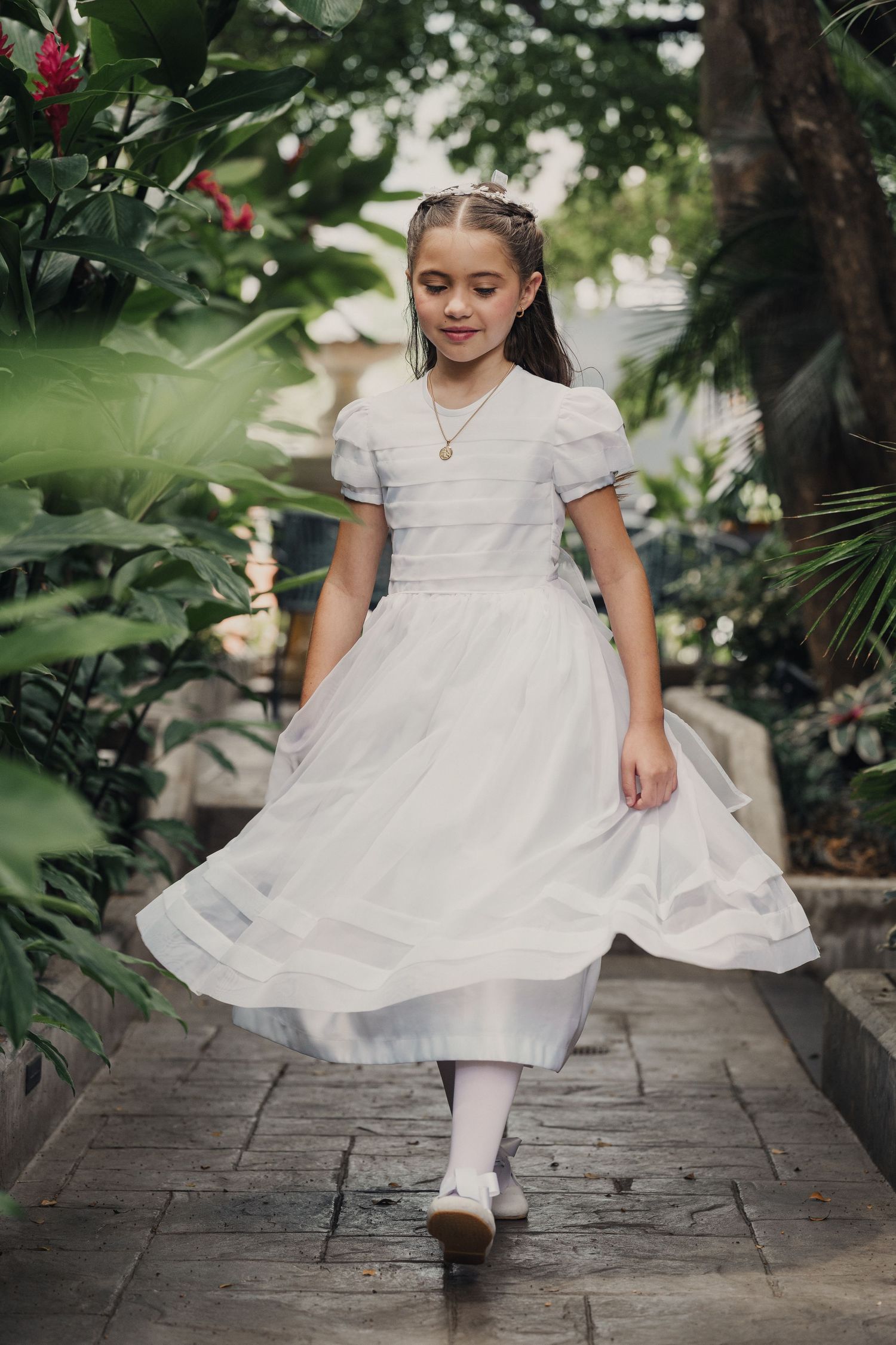 A child in a white formal dress twirls on a stone pathway surrounded by lush greenery and trees.