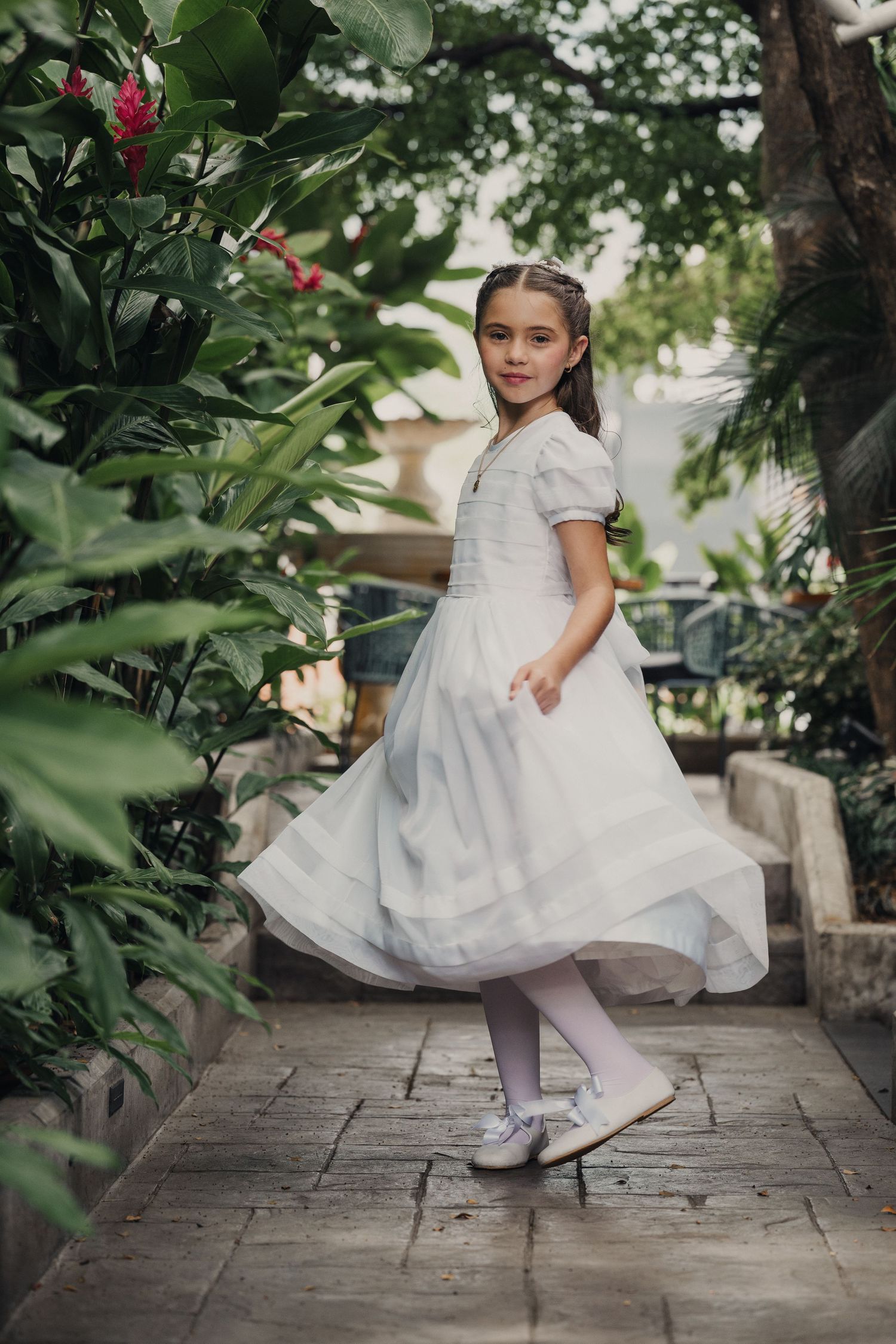 A young girl in a white dress and tiara dances through a garden setting with stone pavers and tropical plants.