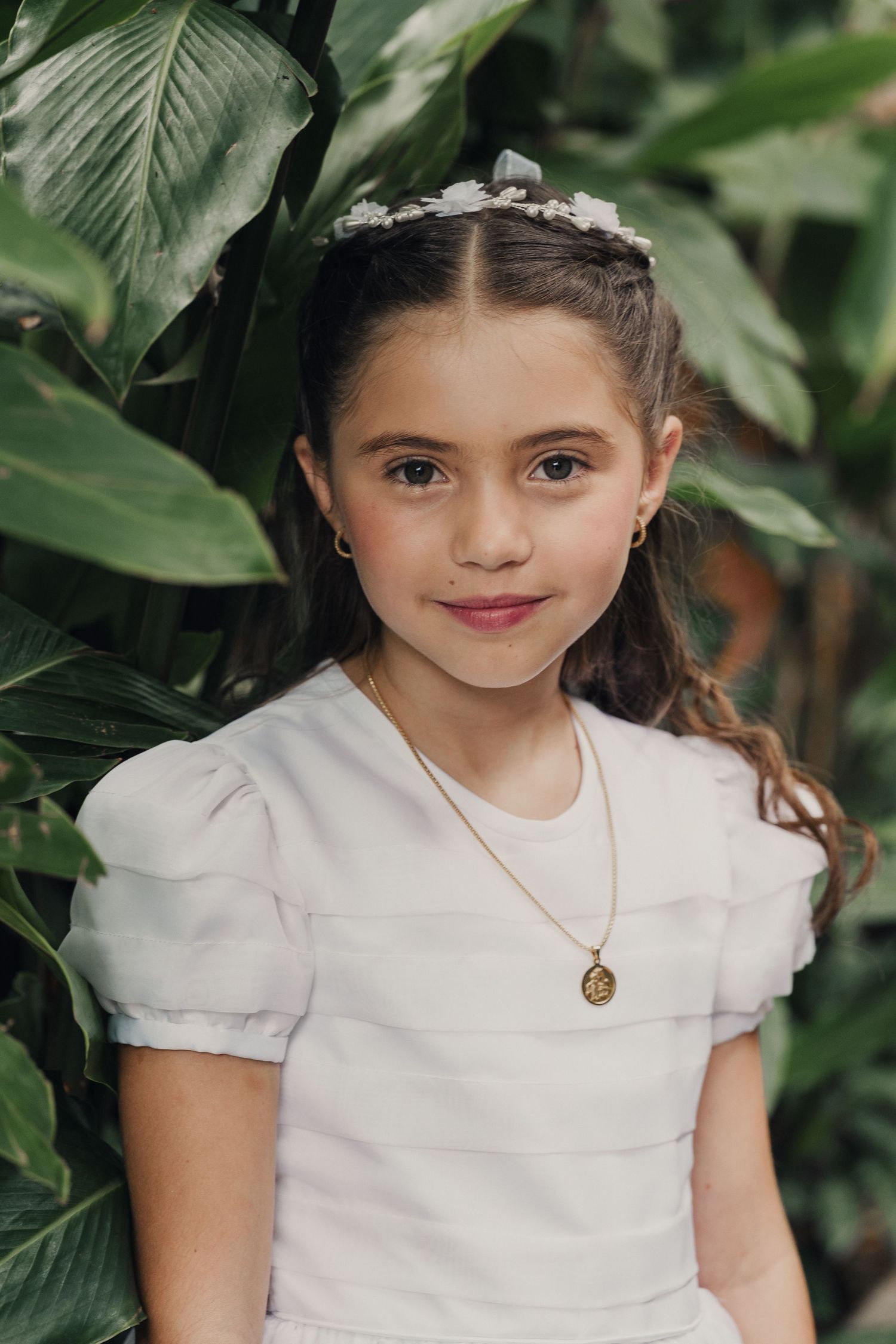 A young person in a white dress poses against lush green foliage in an outdoor portrait setting.