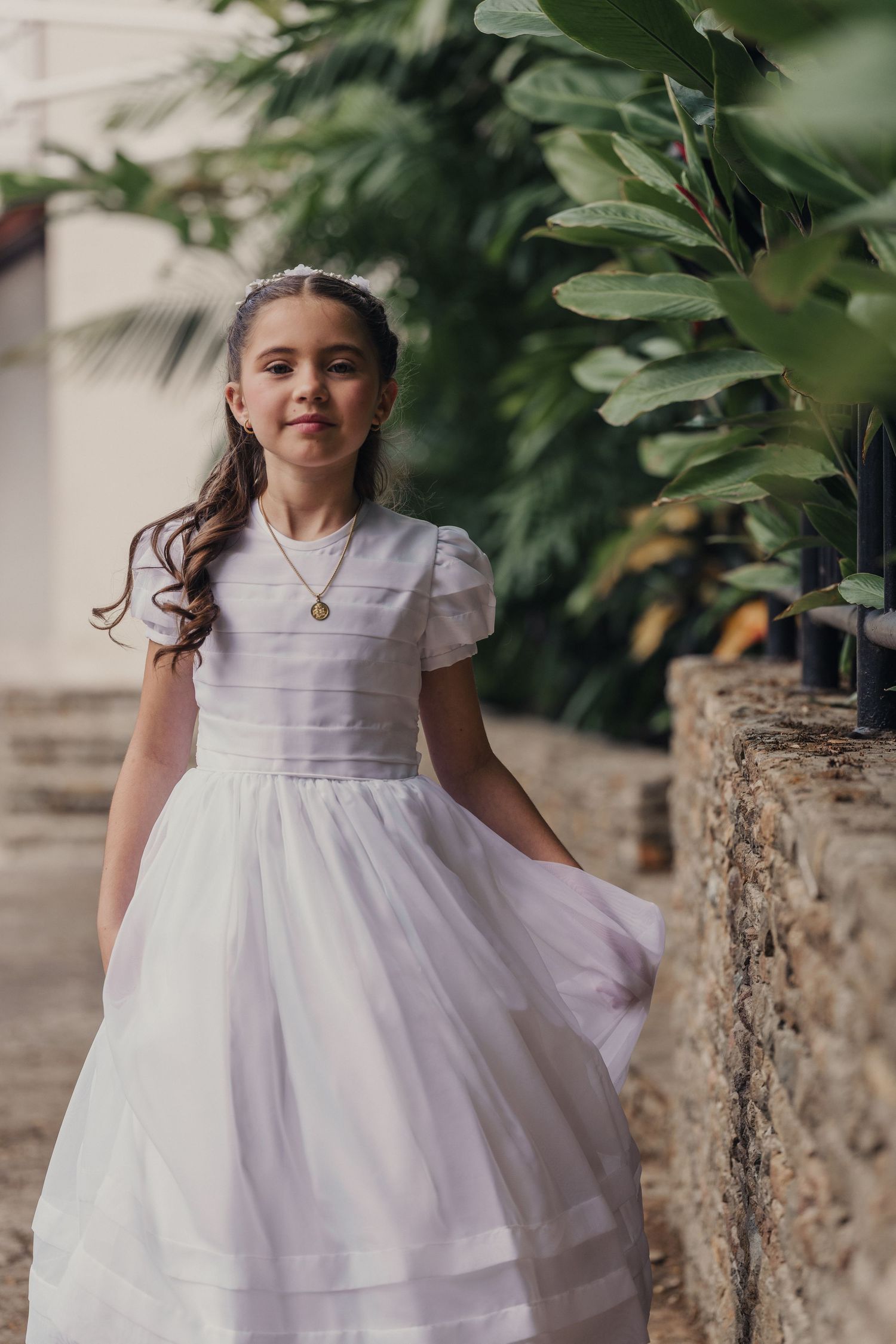 Young girl in white communion or flower girl dress twirls and smiles in garden setting with stone wall backdrop.