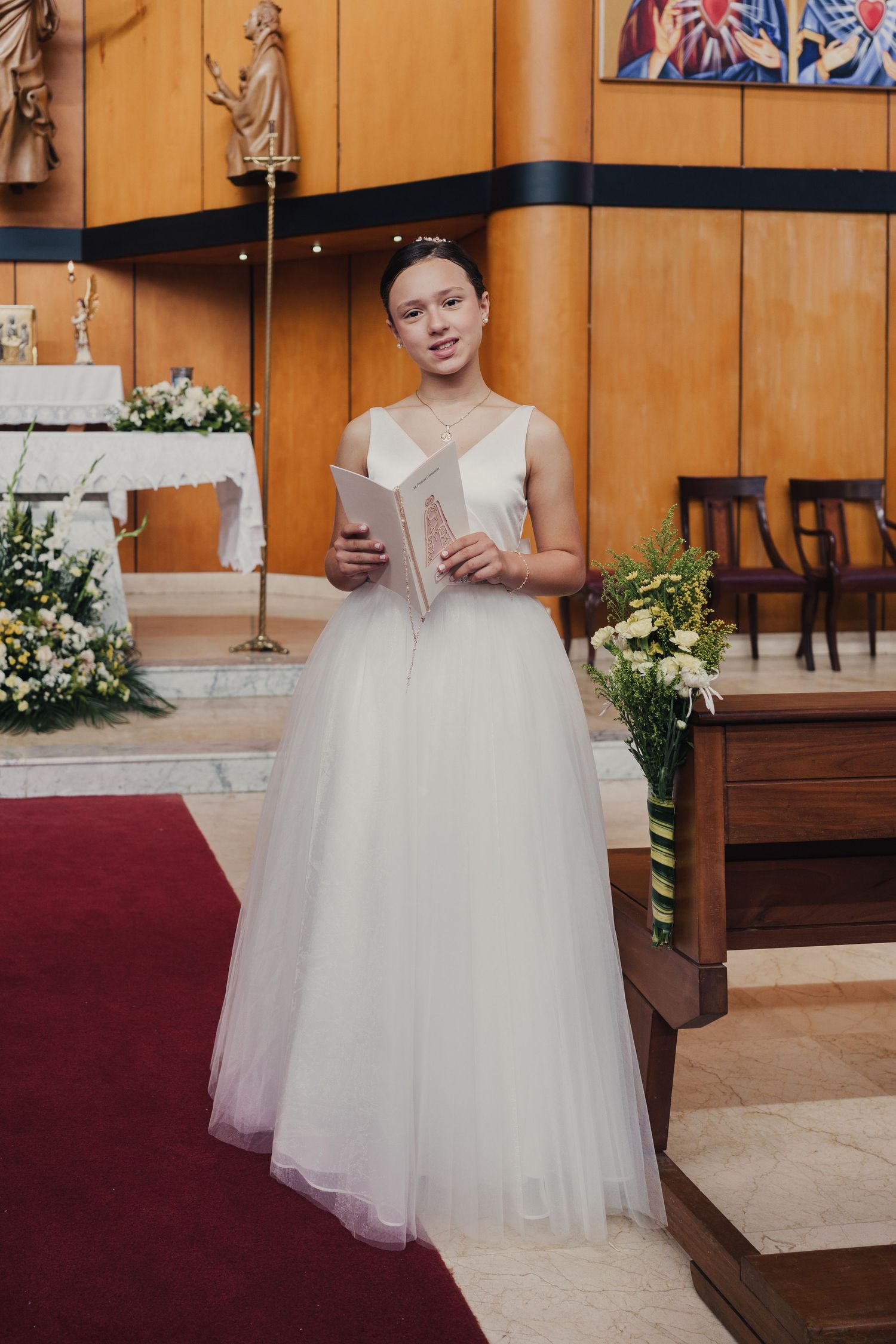 A bride in a white tulle ballgown sits on a wooden church pew during a wedding ceremony.
