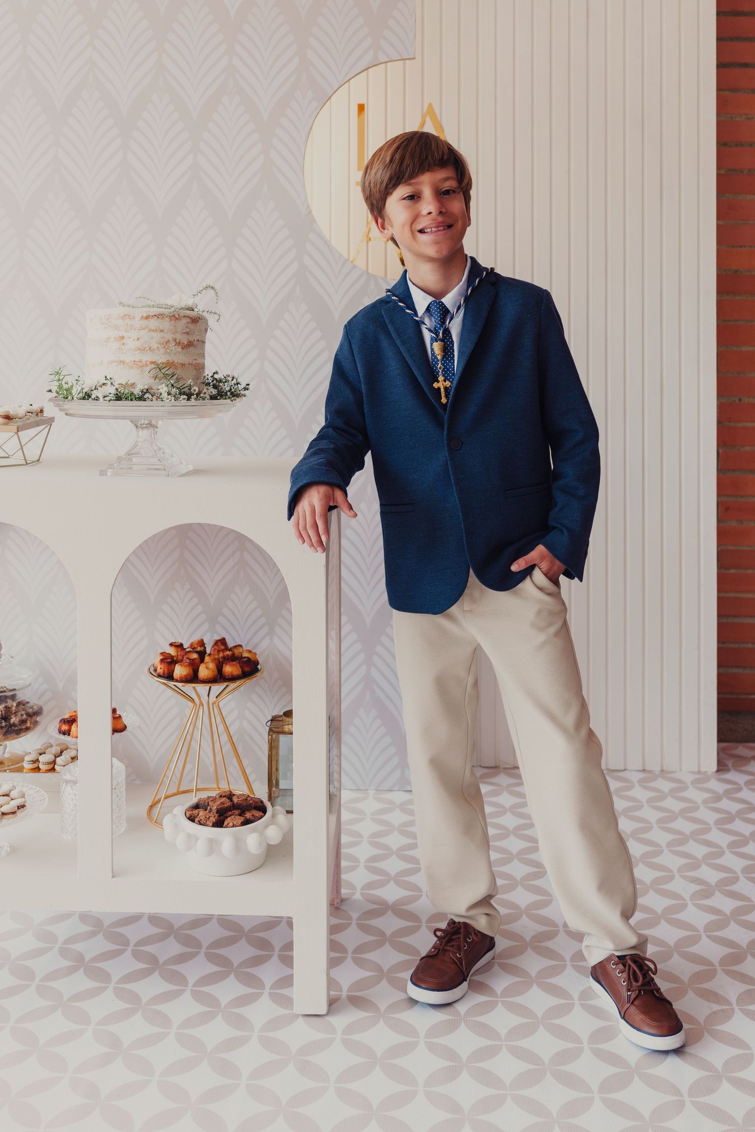 Child in navy blazer and khakis poses beside white geometric display setup with desserts.