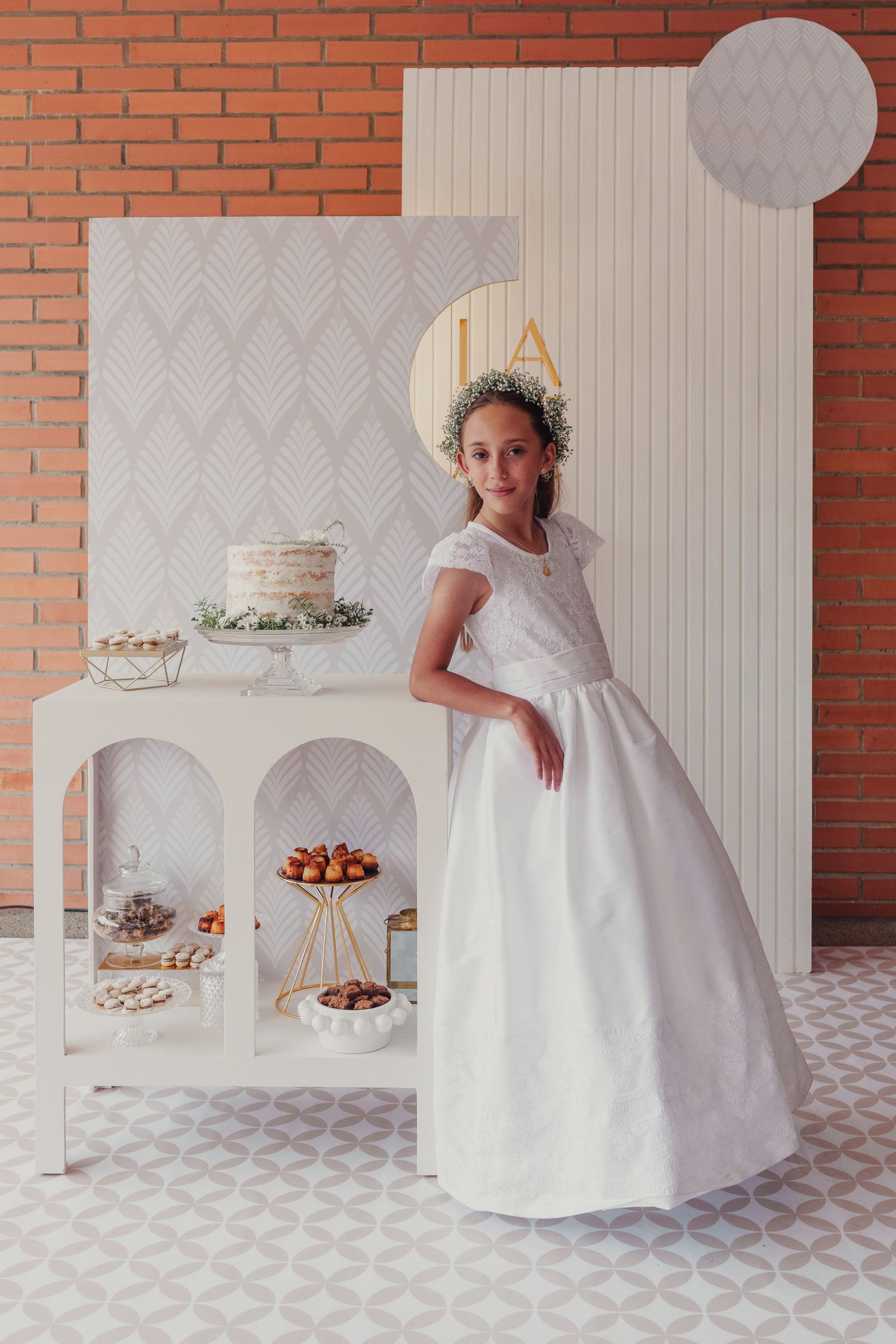 White communion or flower girl dress displayed in elegant party setting with dessert table against brick wall backdrop.