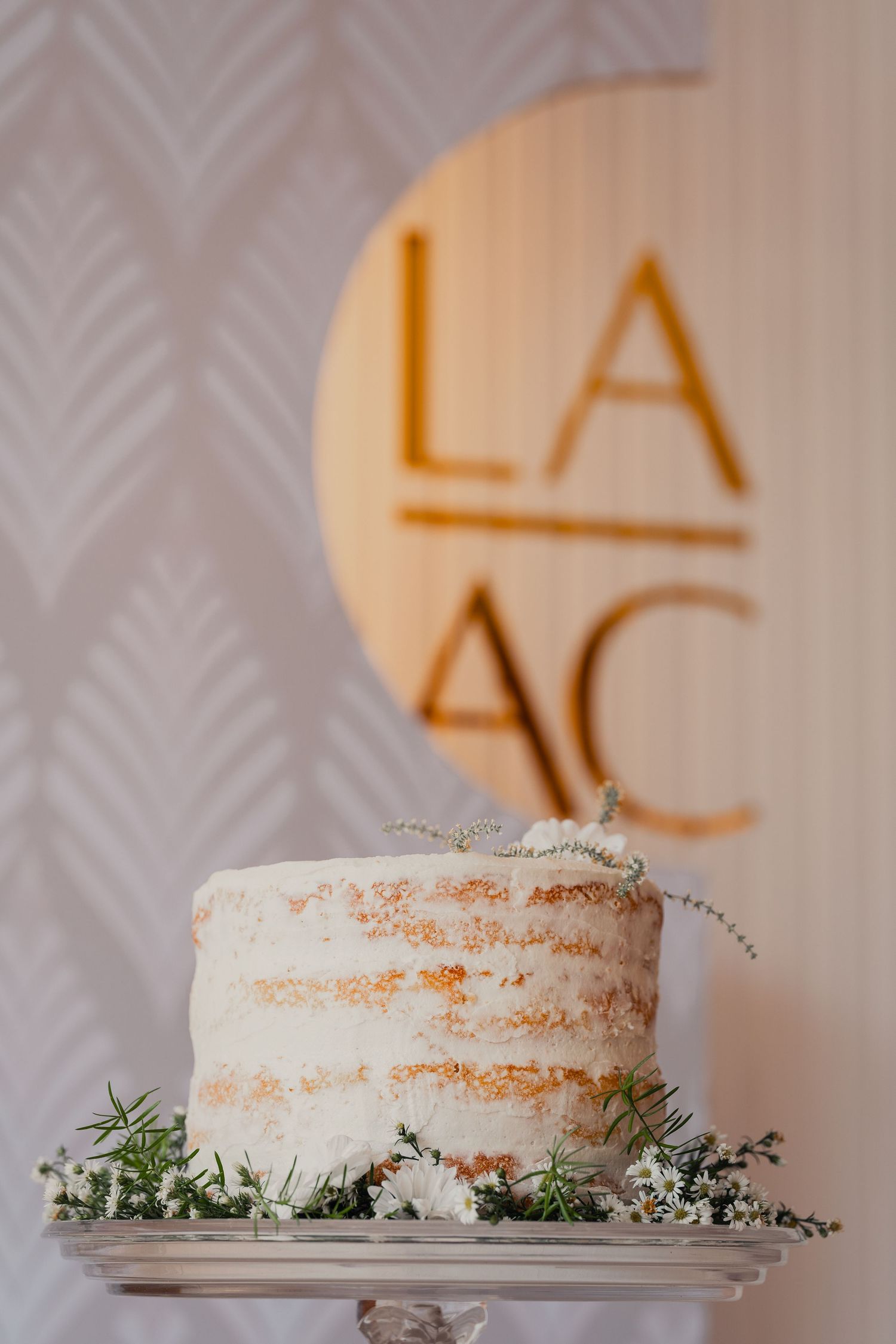 White frosted wedding cake with delicate floral decorations and gold lettering backdrop at reception venue.