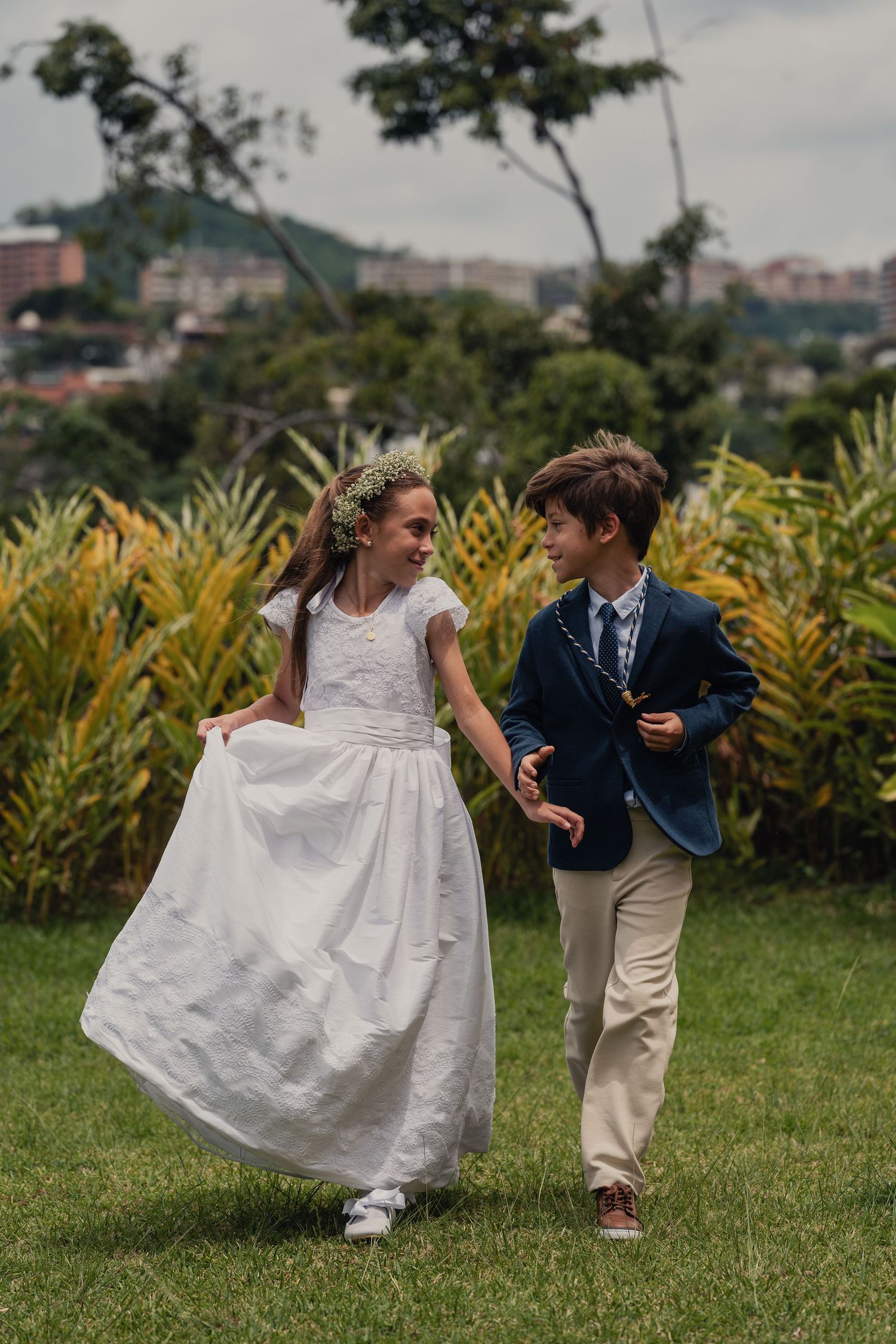 Two children in formal attire walk together on grass, with a girl in white dress and boy in blue blazer and khakis.
