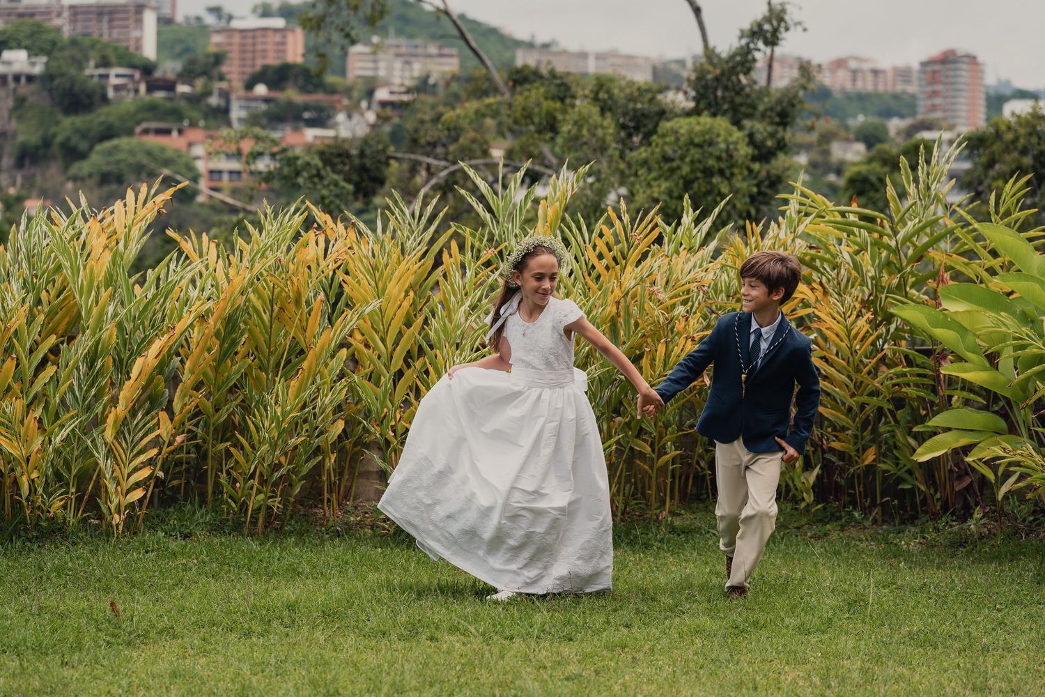 A couple in wedding attire runs joyfully through a lush garden with tall tropical plants in the background.