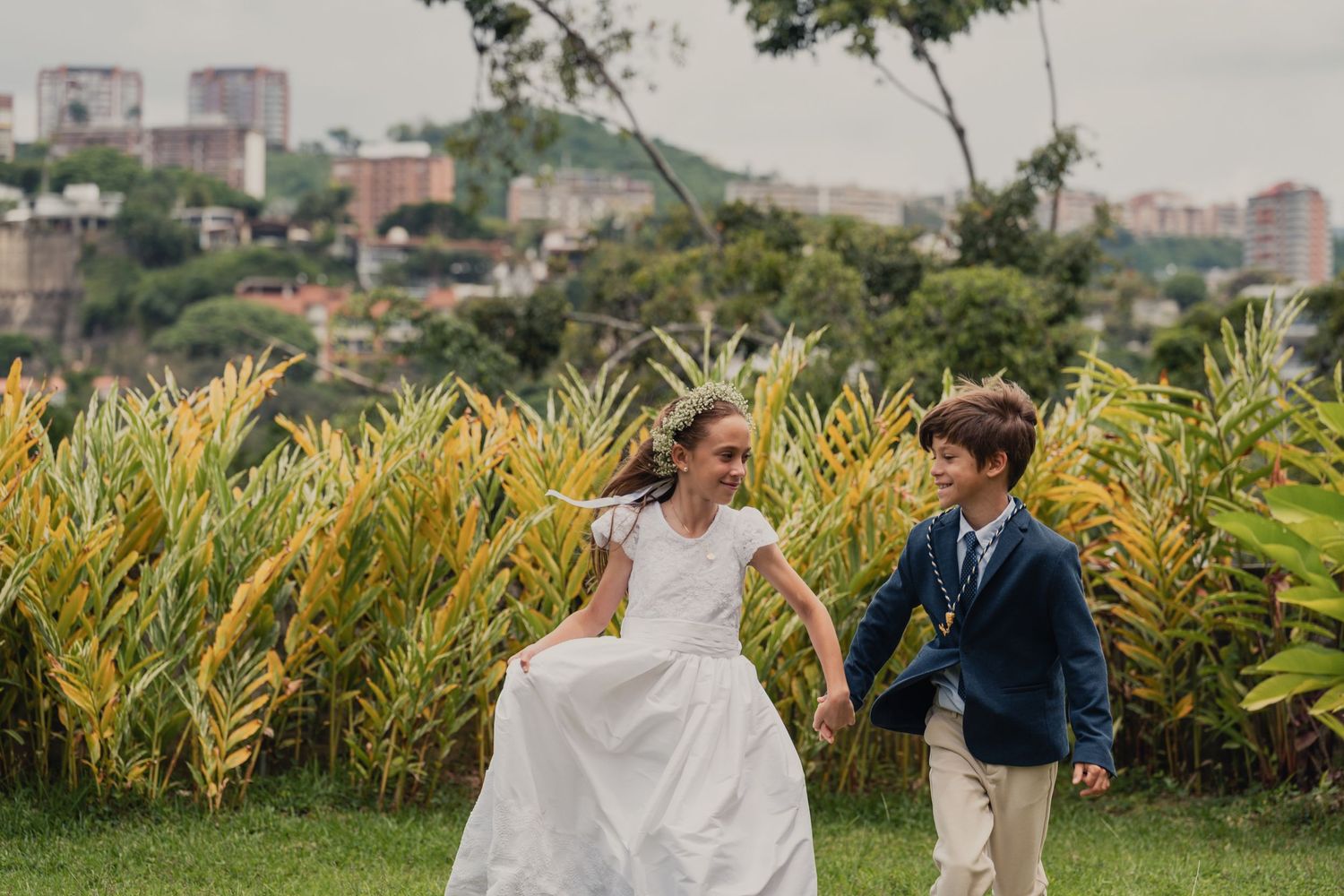 A couple in wedding attire runs joyfully through a garden with yellow plants and city buildings in the background.