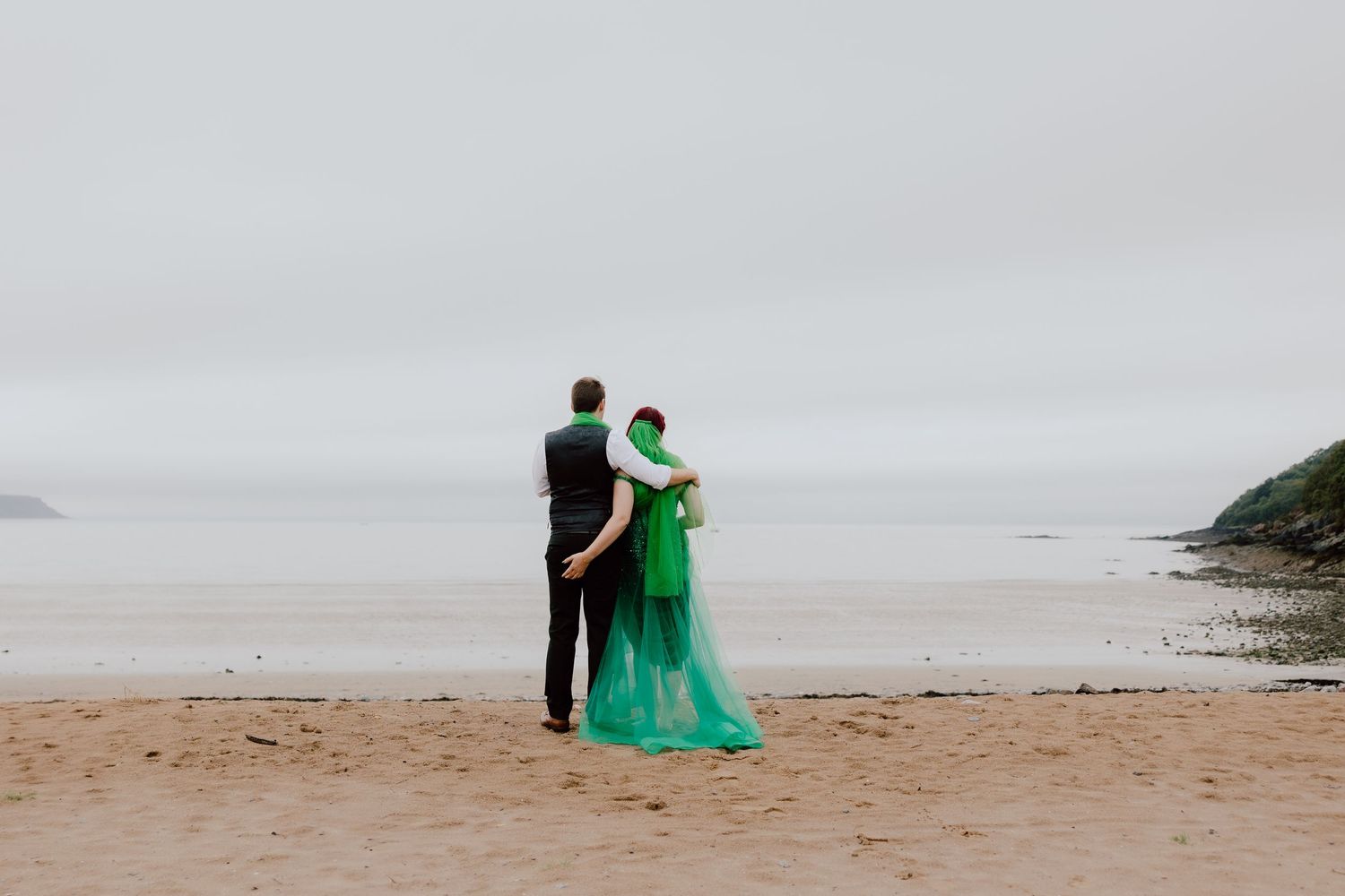 Two figures in black and turquoise attire walk along a misty beach shoreline during an overcast day.