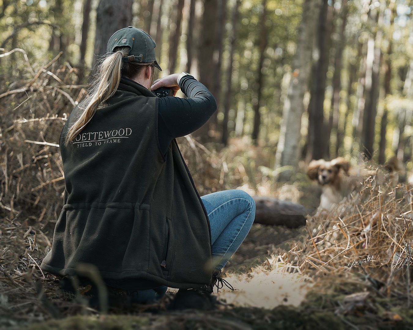 A woman in a green gilet uses a camera in woodland while a spaniel sits nearby.