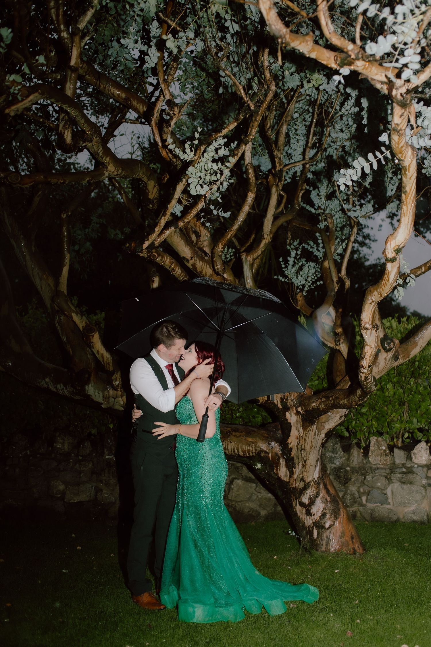 A couple in formal attire embraces under trees at dusk, with the person in a flowing emerald green gown.