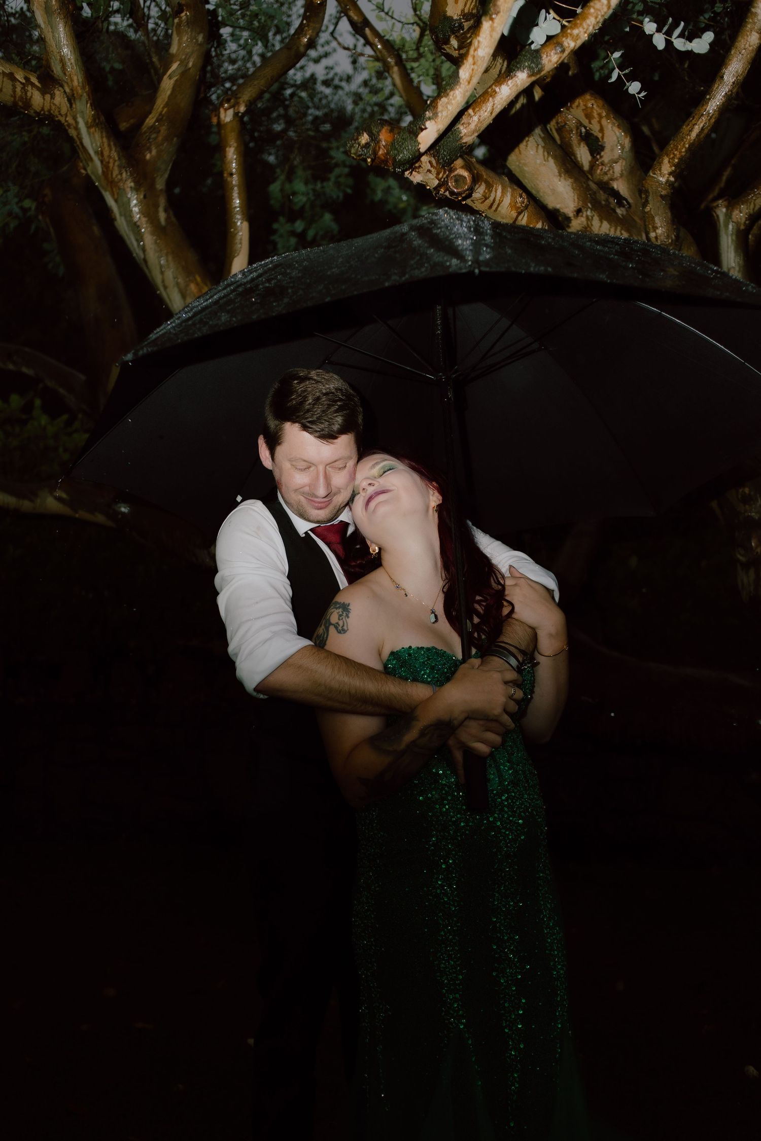 A romantic couple in formal attire share an intimate kiss in a dimly lit garden setting with trees in the background.