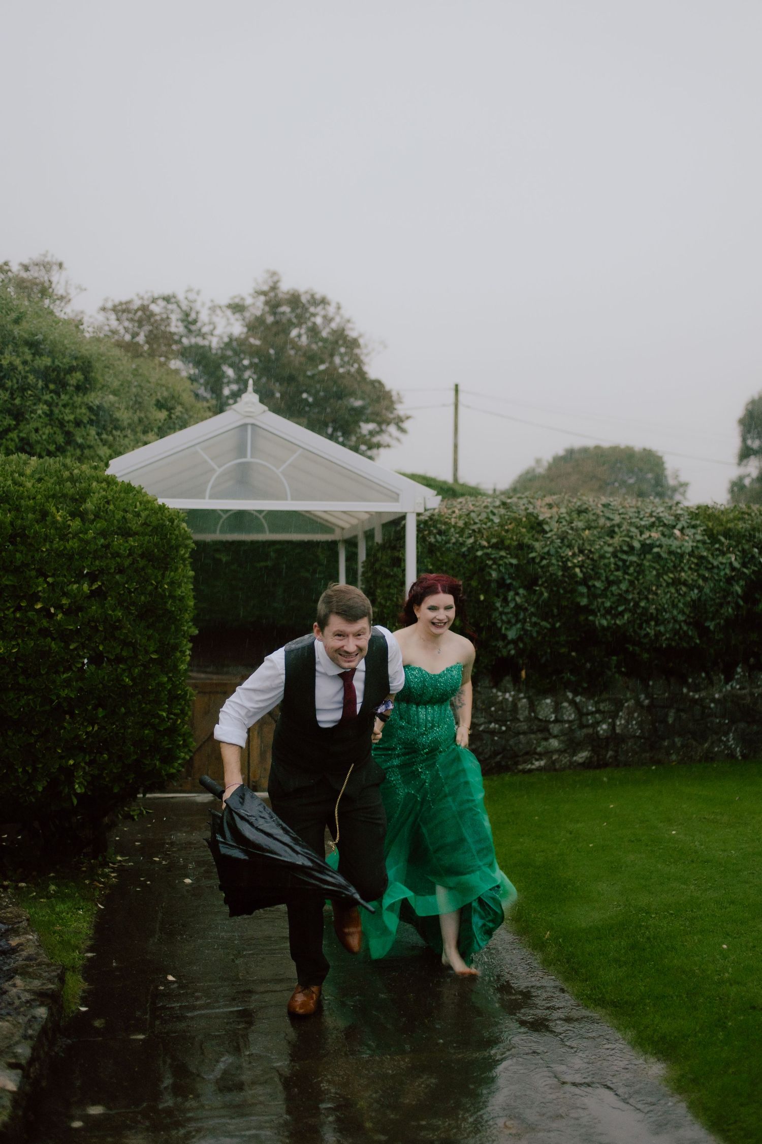 A couple in formal attire playfully runs through the rain on a green lawn.