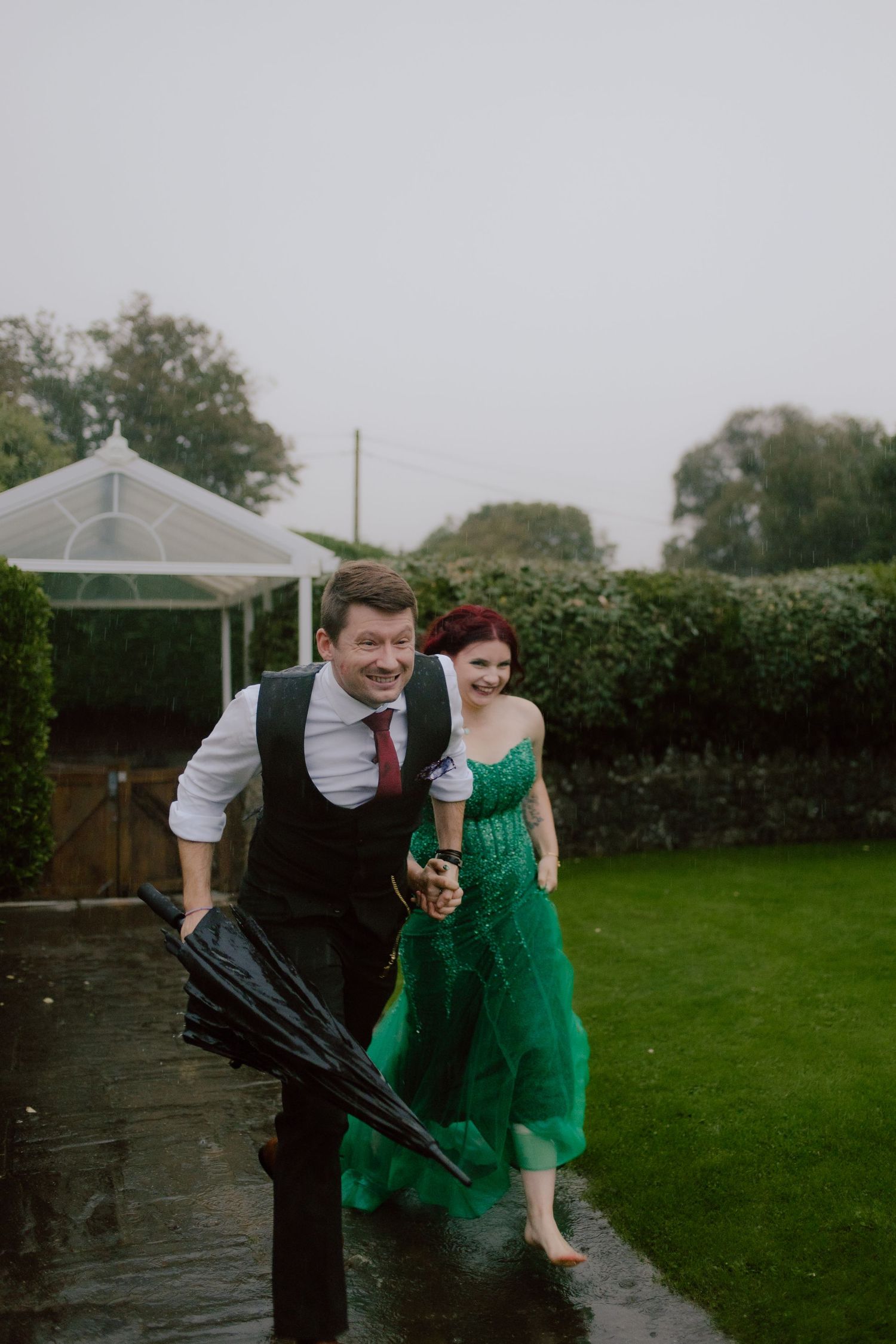 Couple in formal attire playfully poses in the rain under a gazebo in a backyard garden setting.