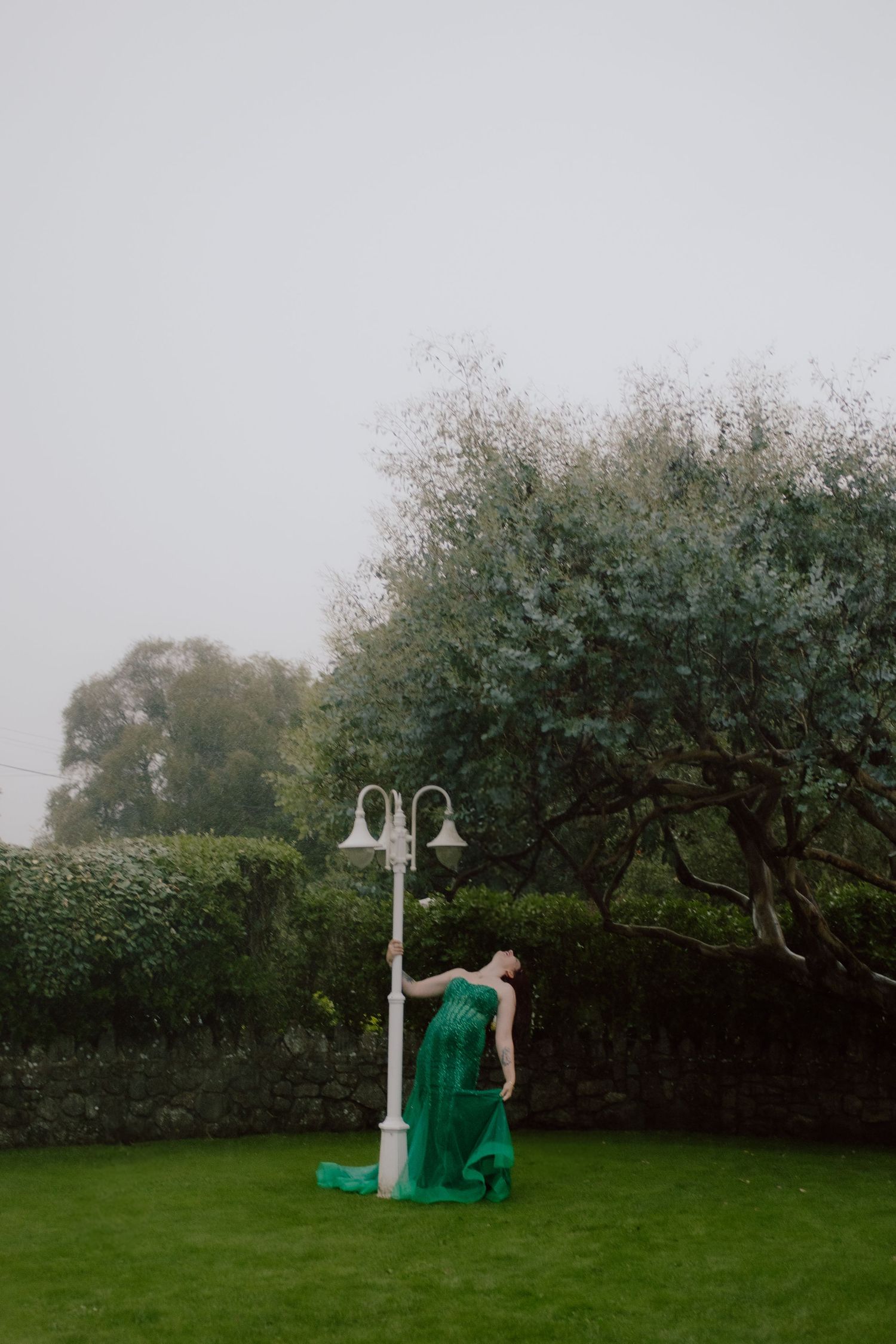 A figure in a flowing emerald green dress poses dramatically near a lamp post on a misty garden lawn.