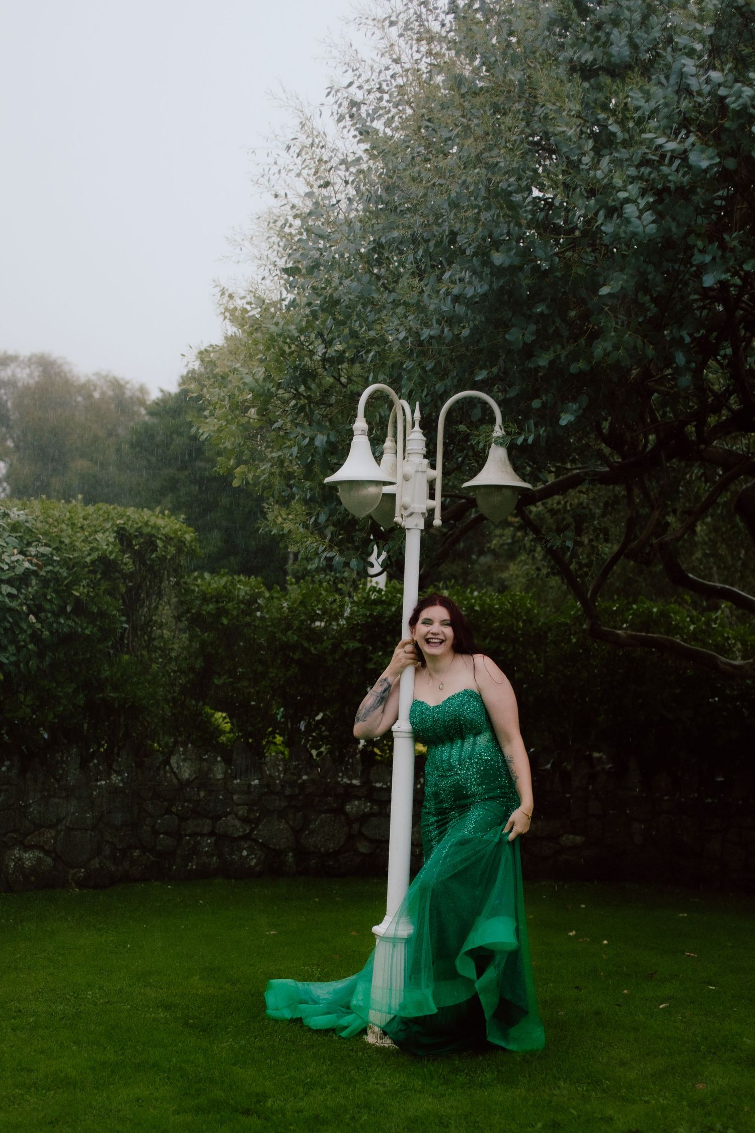 A person in an emerald green dress poses gracefully near a white lamp post in a lush garden setting.