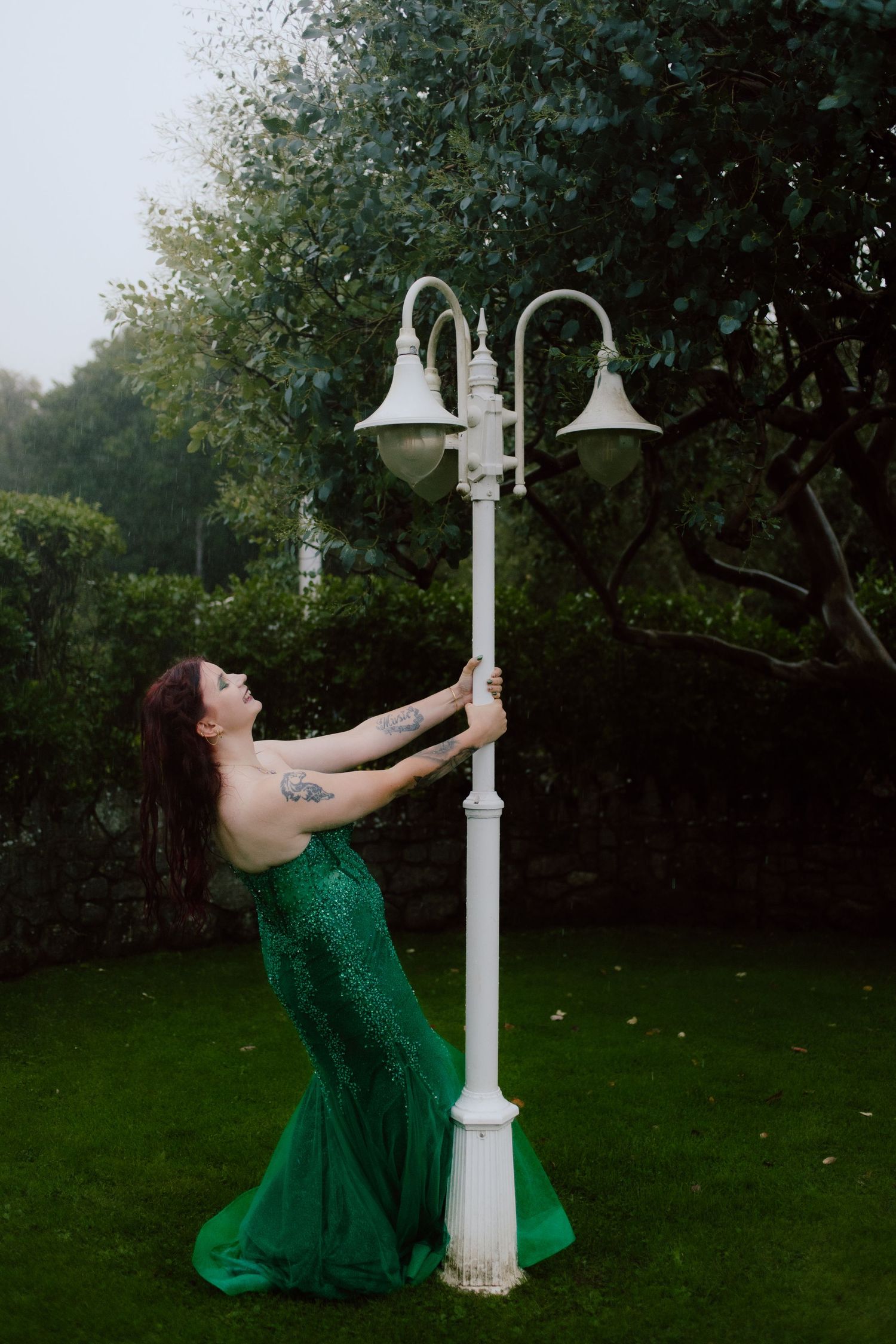 A person in an emerald green dress poses dramatically with a white vintage-style lamppost in a garden setting.