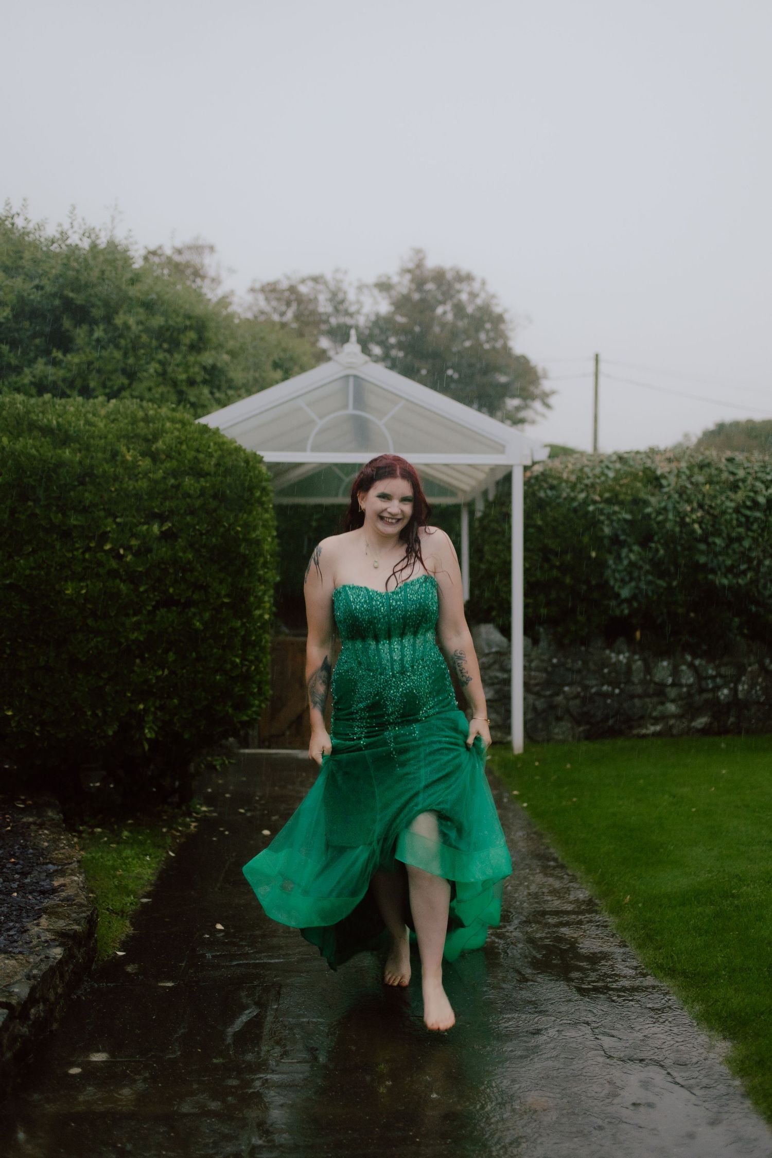 A person in a flowing emerald green dress walks barefoot on a wet garden path beneath a white gazebo.