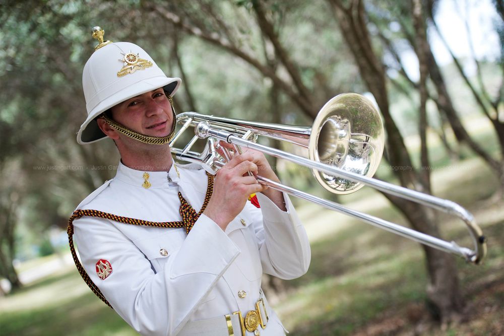 Military band members in white uniforms playing trombones during outdoor performance.