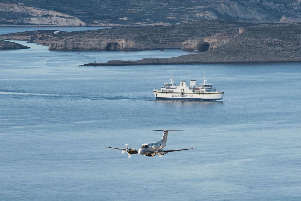 Aircraft and boats traverse a scenic coastal waterway with mountainous landscape in background.