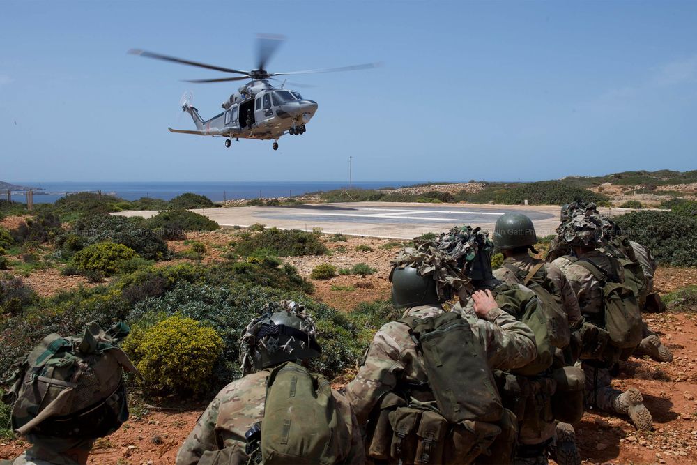 Military helicopter approaching landing zone with troops in defensive positions on rocky terrain.