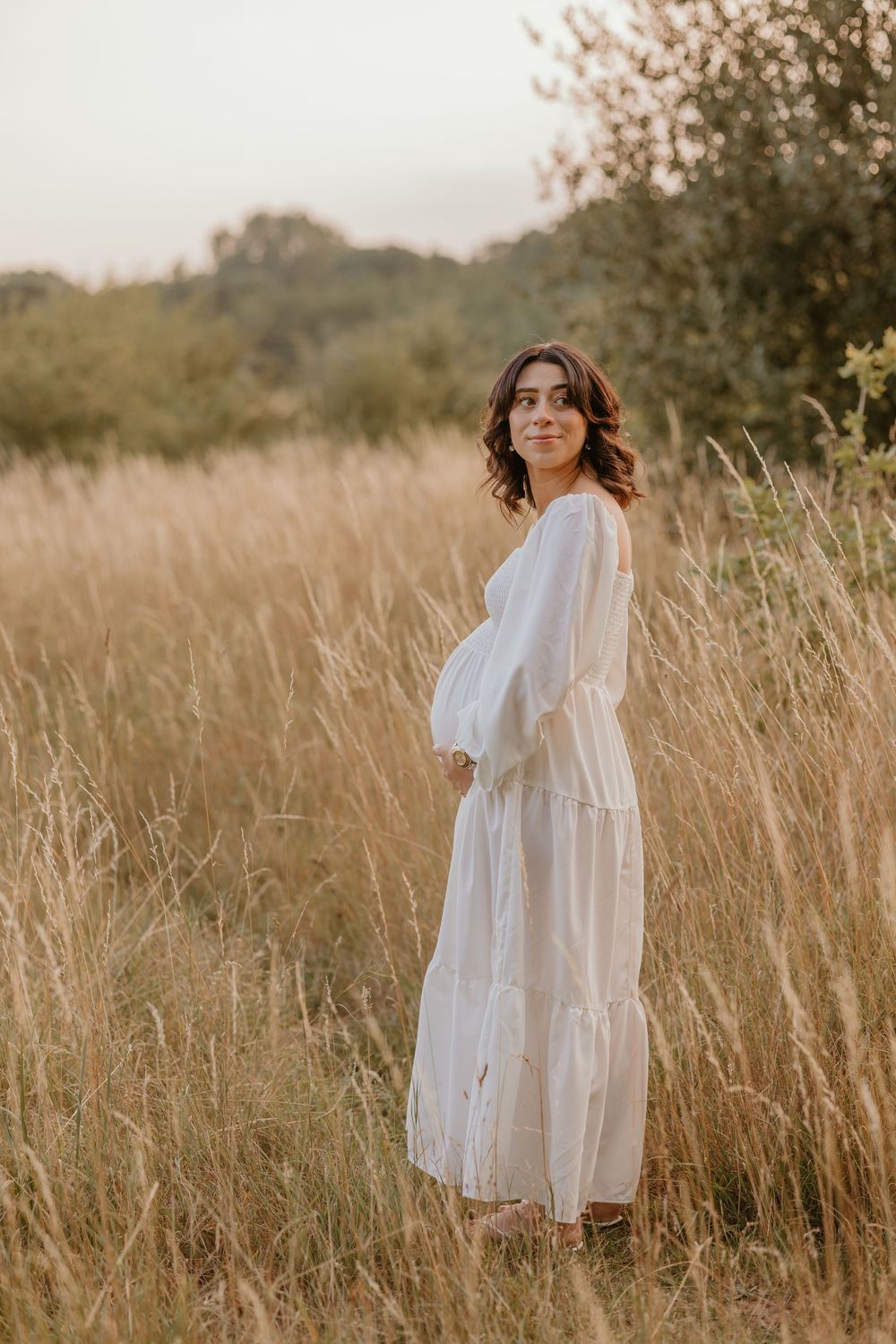 Expectant mother in flowing white dress poses in golden field at sunset.