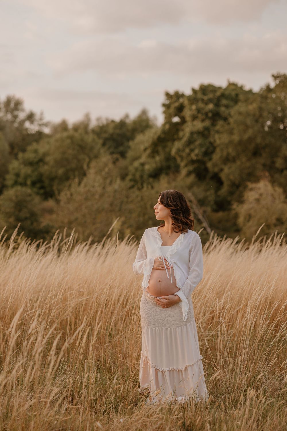 Pregnant woman in white dress standing thoughtfully in tall autumn grass.