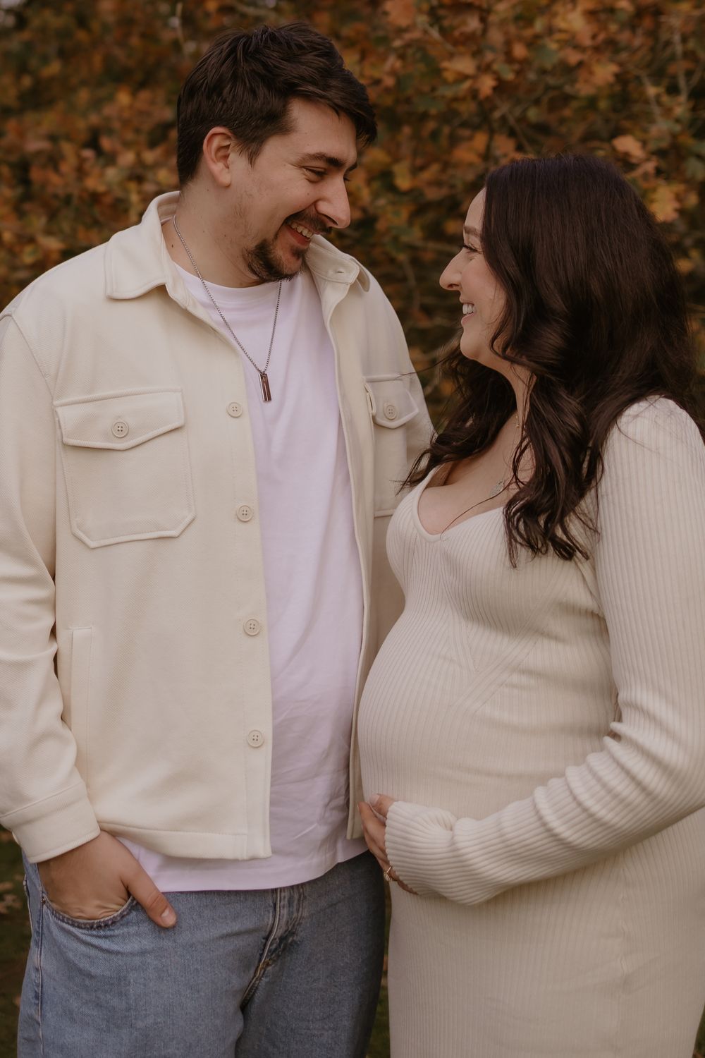 Casual maternity portrait of couple sharing a laugh against autumn foliage.