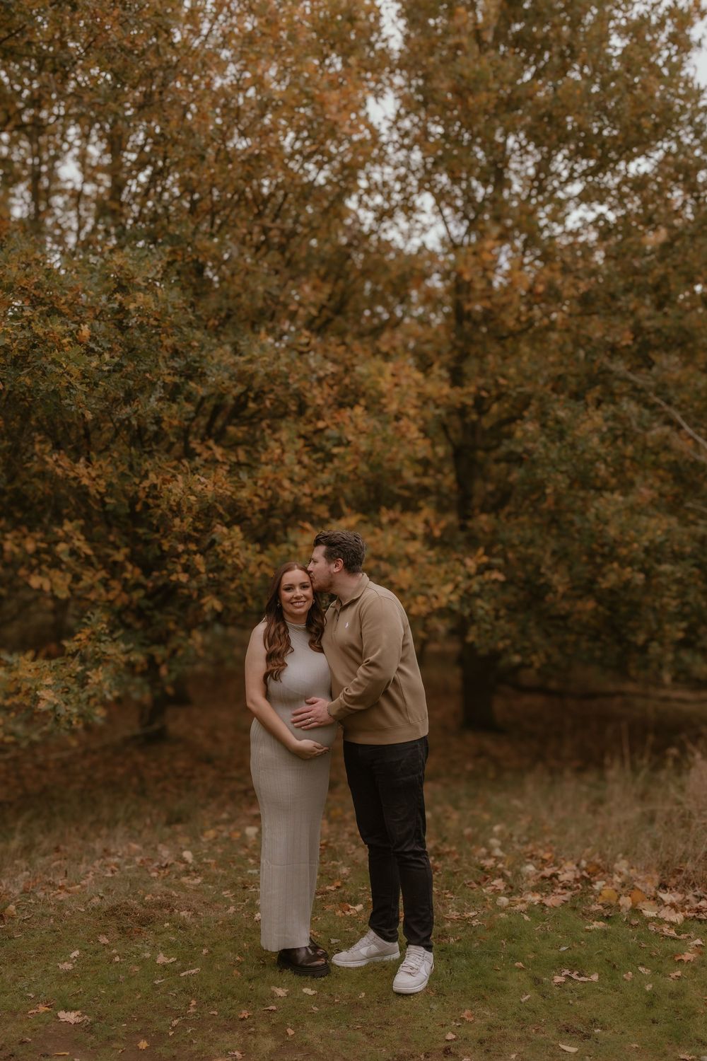 Autumn maternity photoshoot with couple standing together among orange-leaved trees.