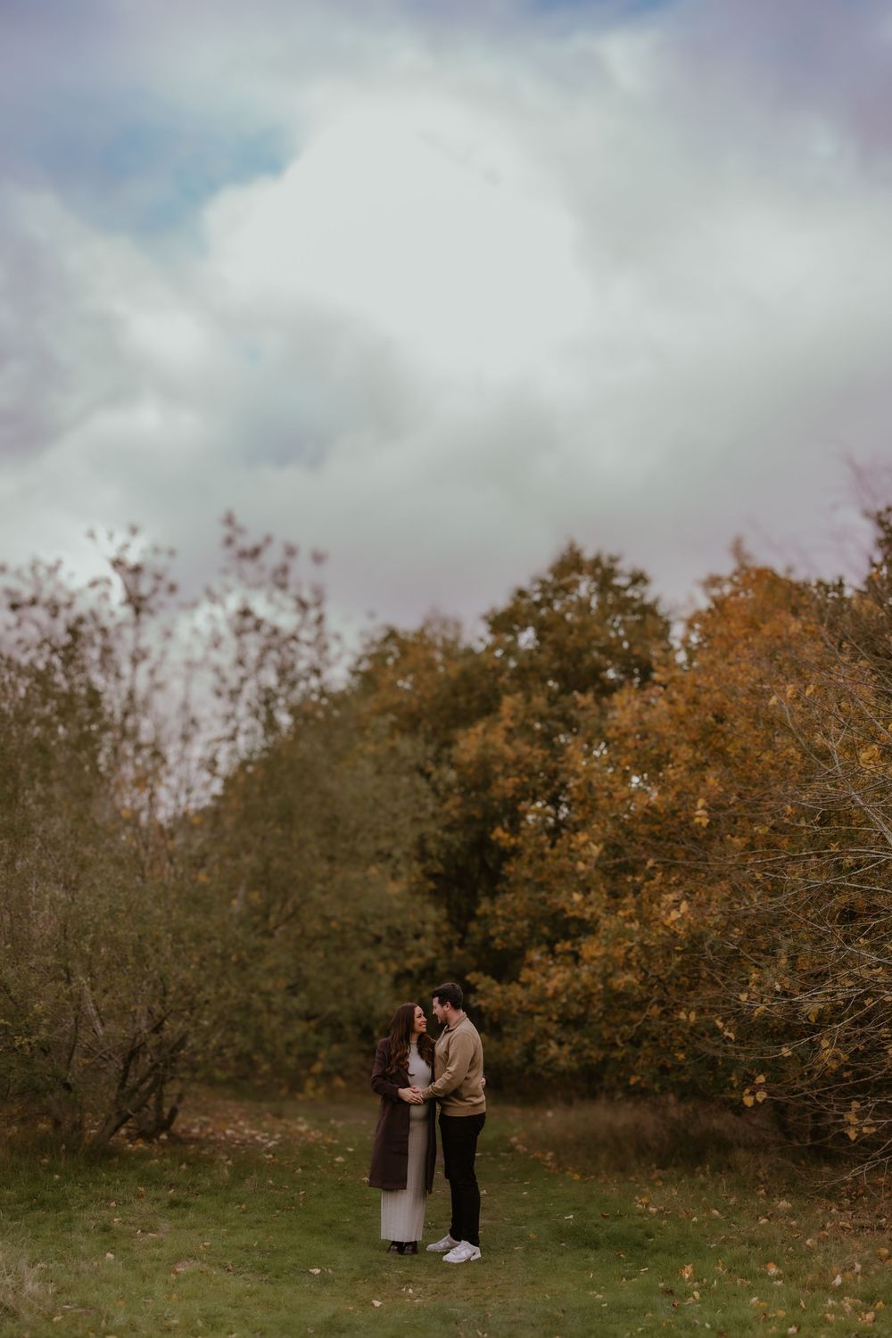 Distant shot of couple embracing under cloudy sky with fall trees in background.