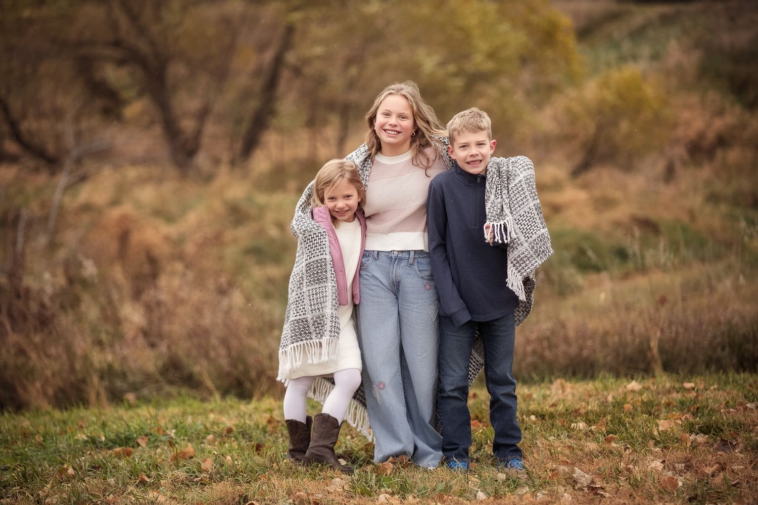 A family group stands together outdoors in autumn, wrapped in cozy blankets against a backdrop of fall foliage.