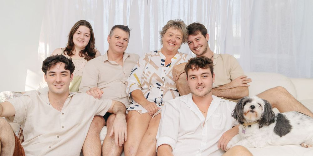 A group in light colored clothing sits together on a white couch with a small dog.