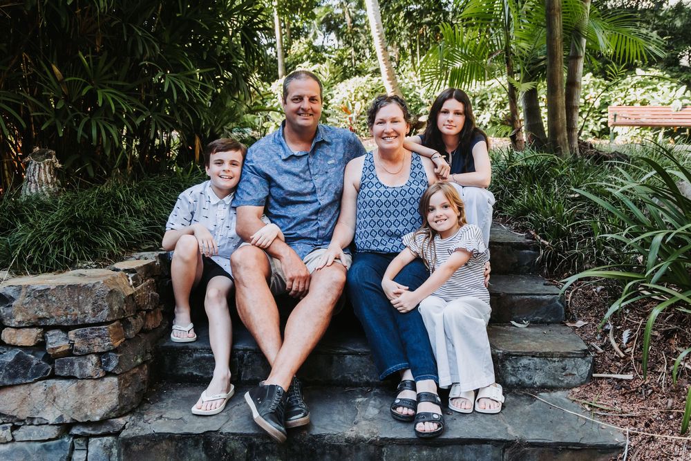 A family sits together on stone steps surrounded by lush tropical landscaping in an outdoor garden setting.