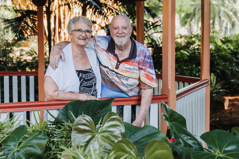 Elderly couple sitting together on a porch swing surrounded by lush green plants and white railings.