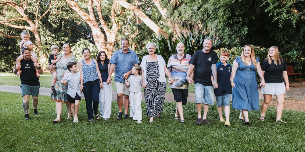 A row of wedding guests in casual attire stand in line outdoors against a lush green garden backdrop.