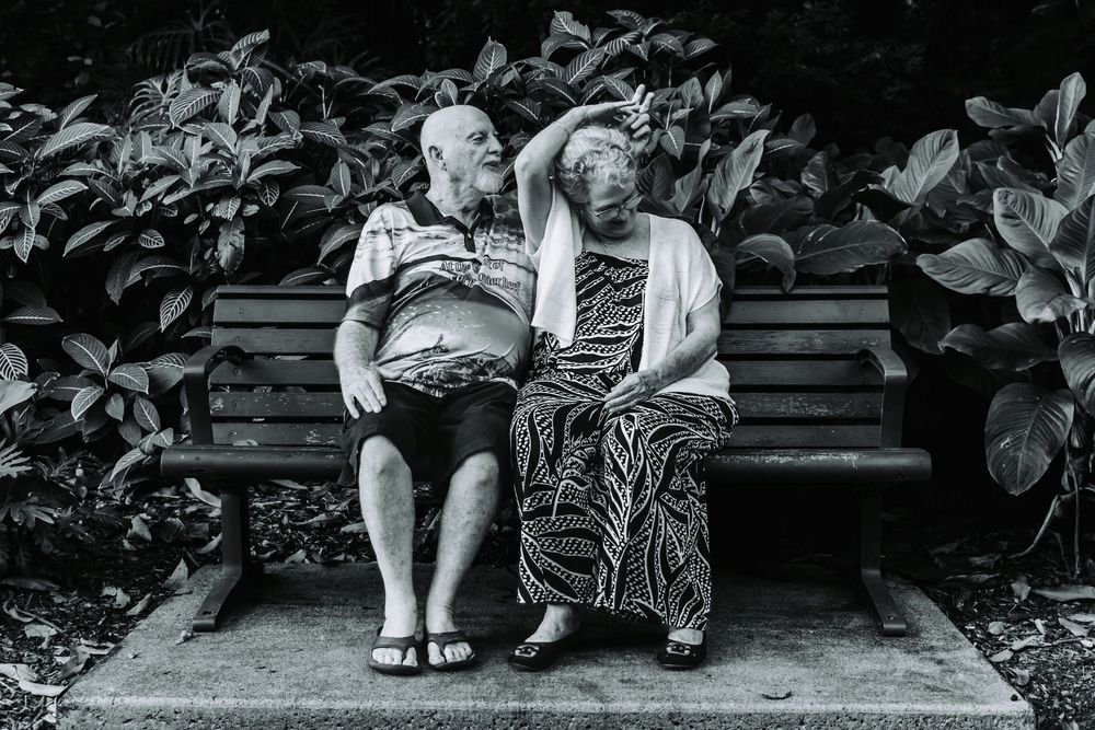 Black and white photo of two people sitting on a wooden bench in a garden setting.