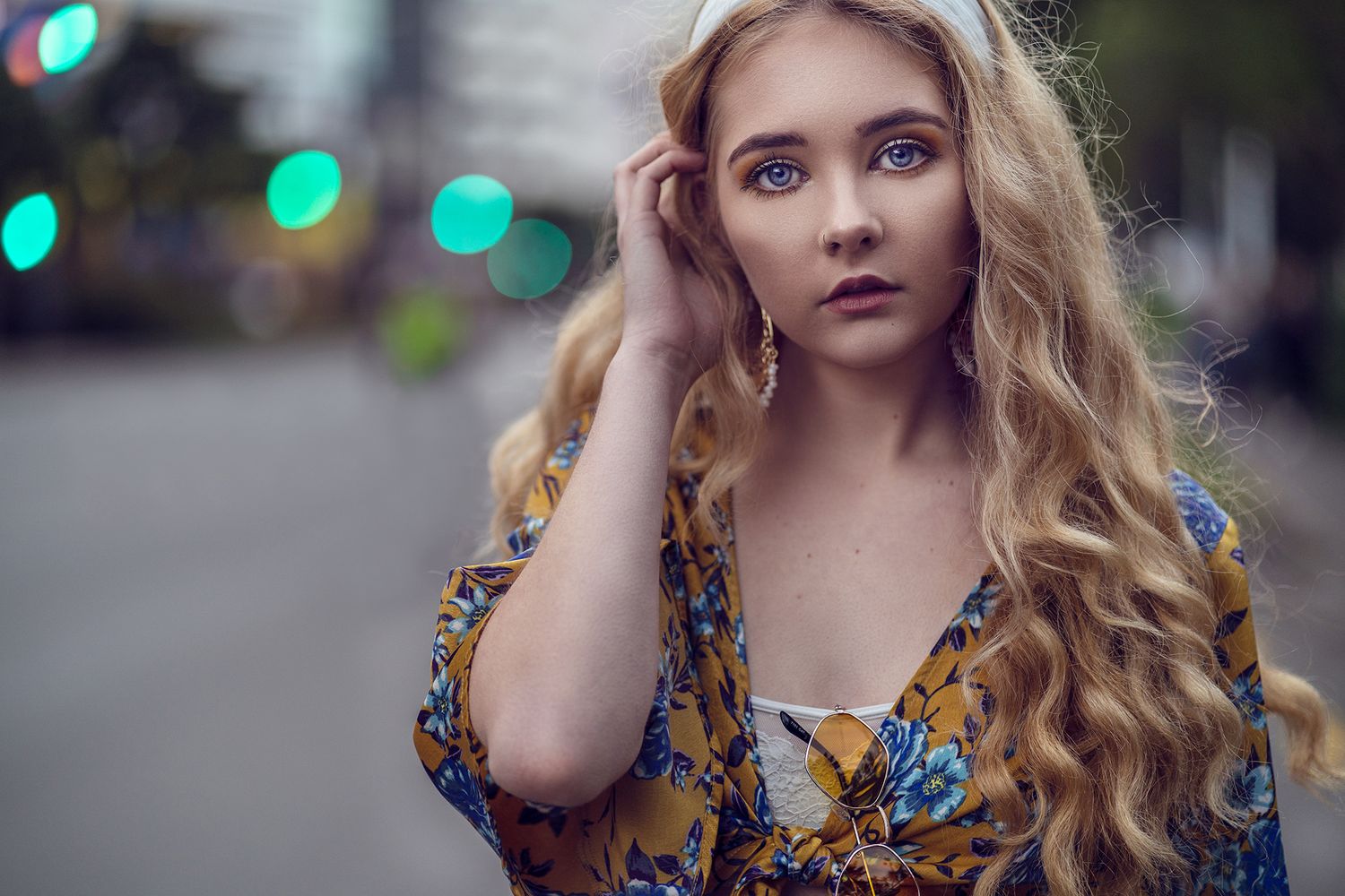 Young woman with long blonde hair in a floral top standing on a city street, holding her hair with blurred streetlights in the background.