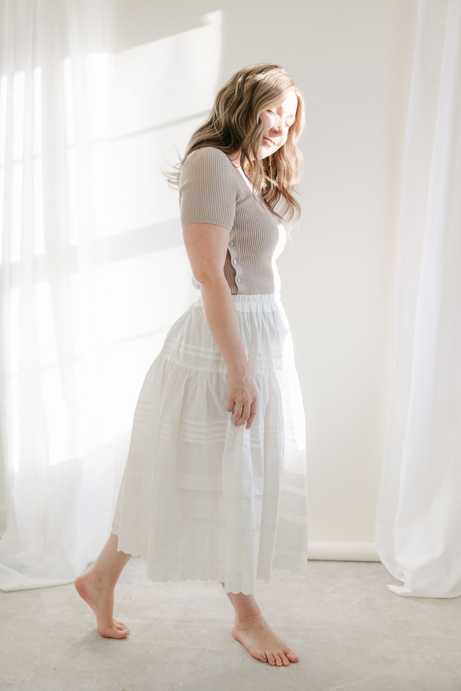 Woman in white dress dances joyfully in bright sunlit room with flowing curtains and bare feet.