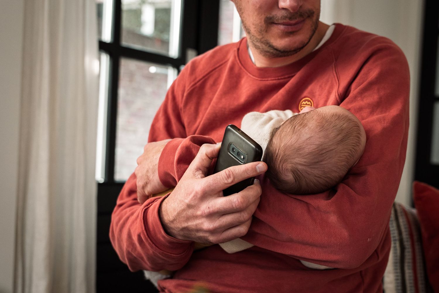 A person in a coral sweatshirt tenderly holds a sleeping newborn baby near a window.