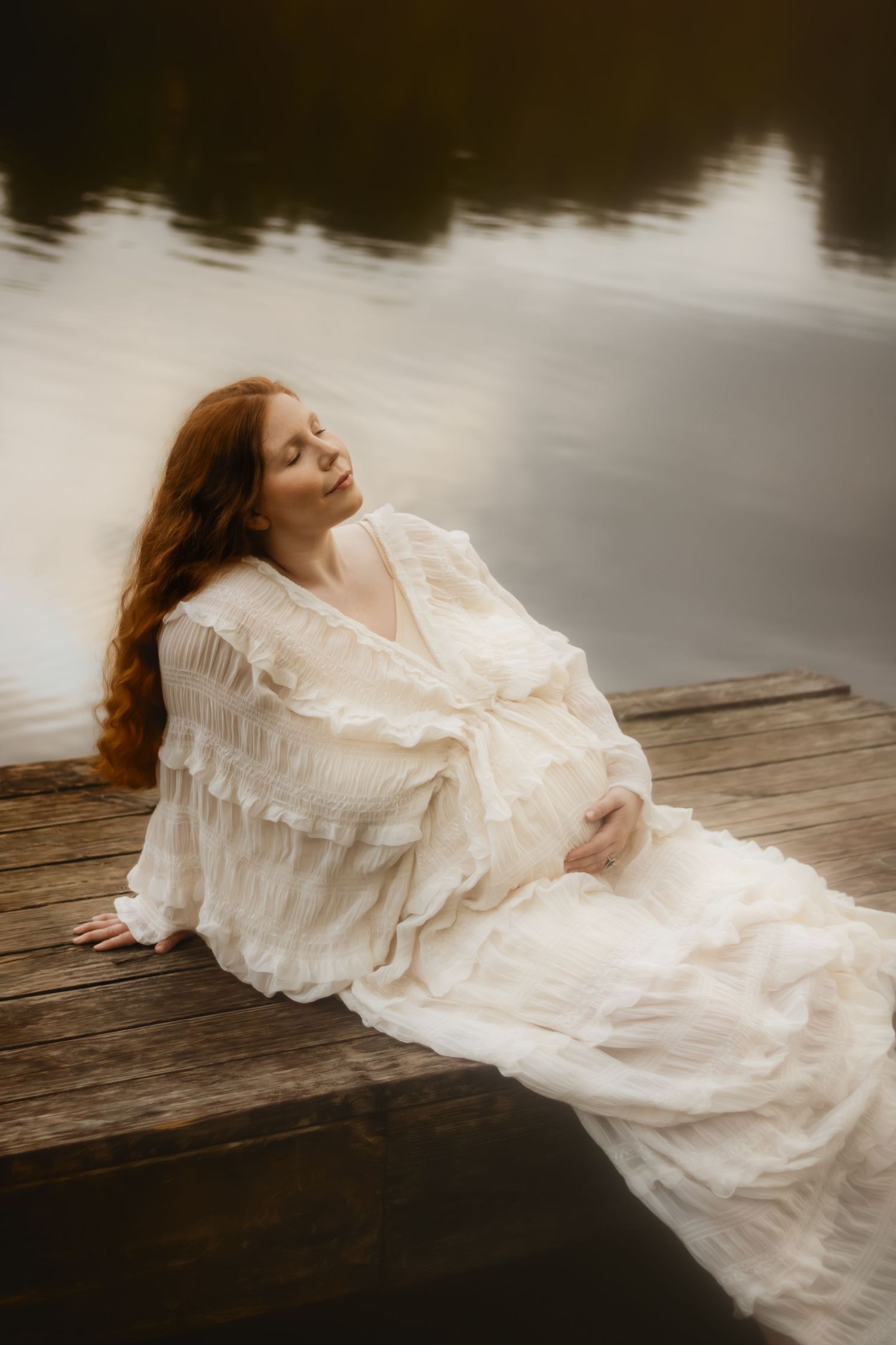 A dreamy maternity portrait of a redheaded woman in a white gown relaxing on a lakeside wooden dock.