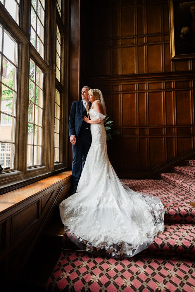 Bride and groom share a romantic moment by a grand window on ornate stairs in an elegant wood-paneled venue.