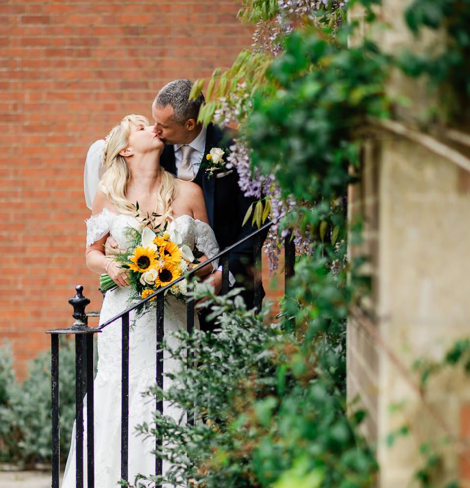 A bride and groom share a romantic kiss on steps surrounded by lush greenery, bride holding a vibrant sunflower bouquet.