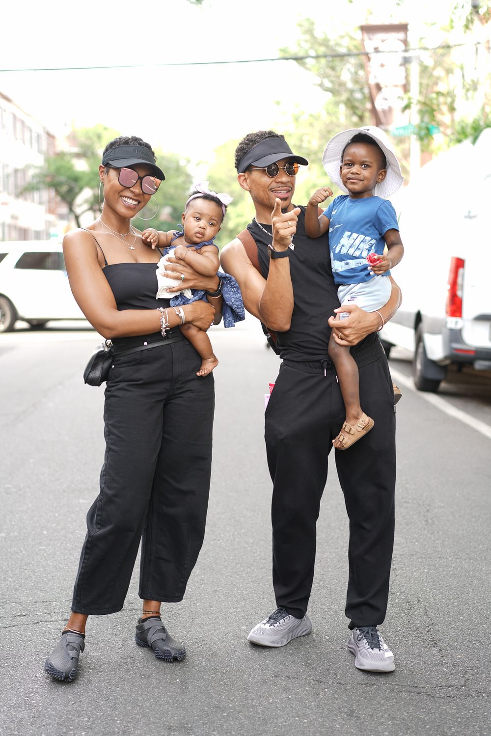 Two women in matching black outfits and visors pose on a street, each holding a baby outdoors.
