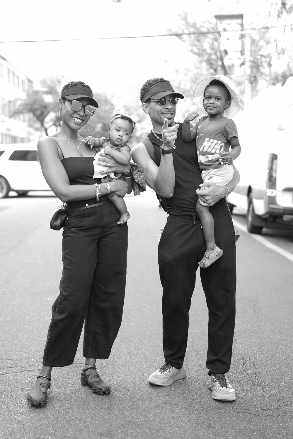 Two adults in matching black outfits and visors hold two babies on a city street in black and white photo.