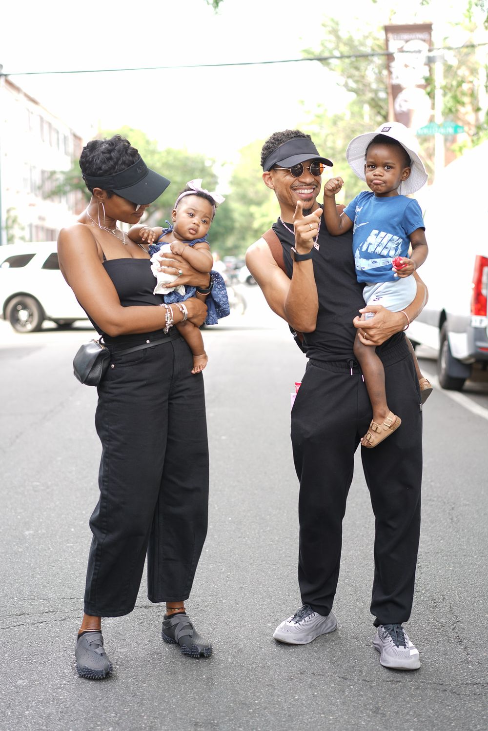 Two women in matching black outfits and visors hold babies on a sunny urban street.