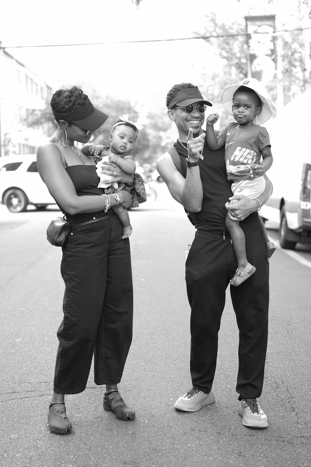 Two stylish adults holding babies on a sunny street, captured in black and white photography.